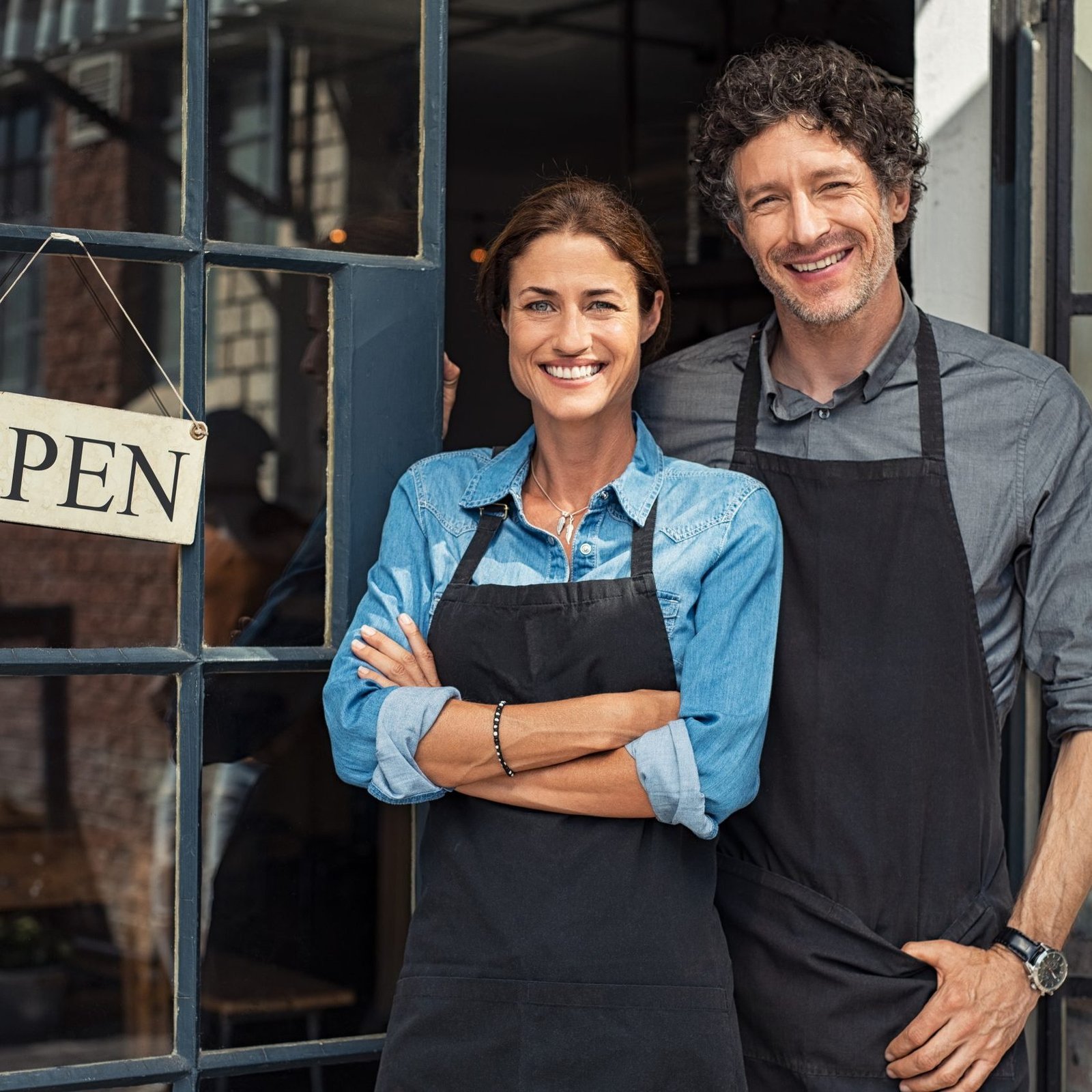 Two cheerful small business owners smiling and looking at camera while standing at entrance door. Happy mature man and mid woman at entrance of newly opened restaurant with open sign board. Smiling couple welcoming customers to small business shop.